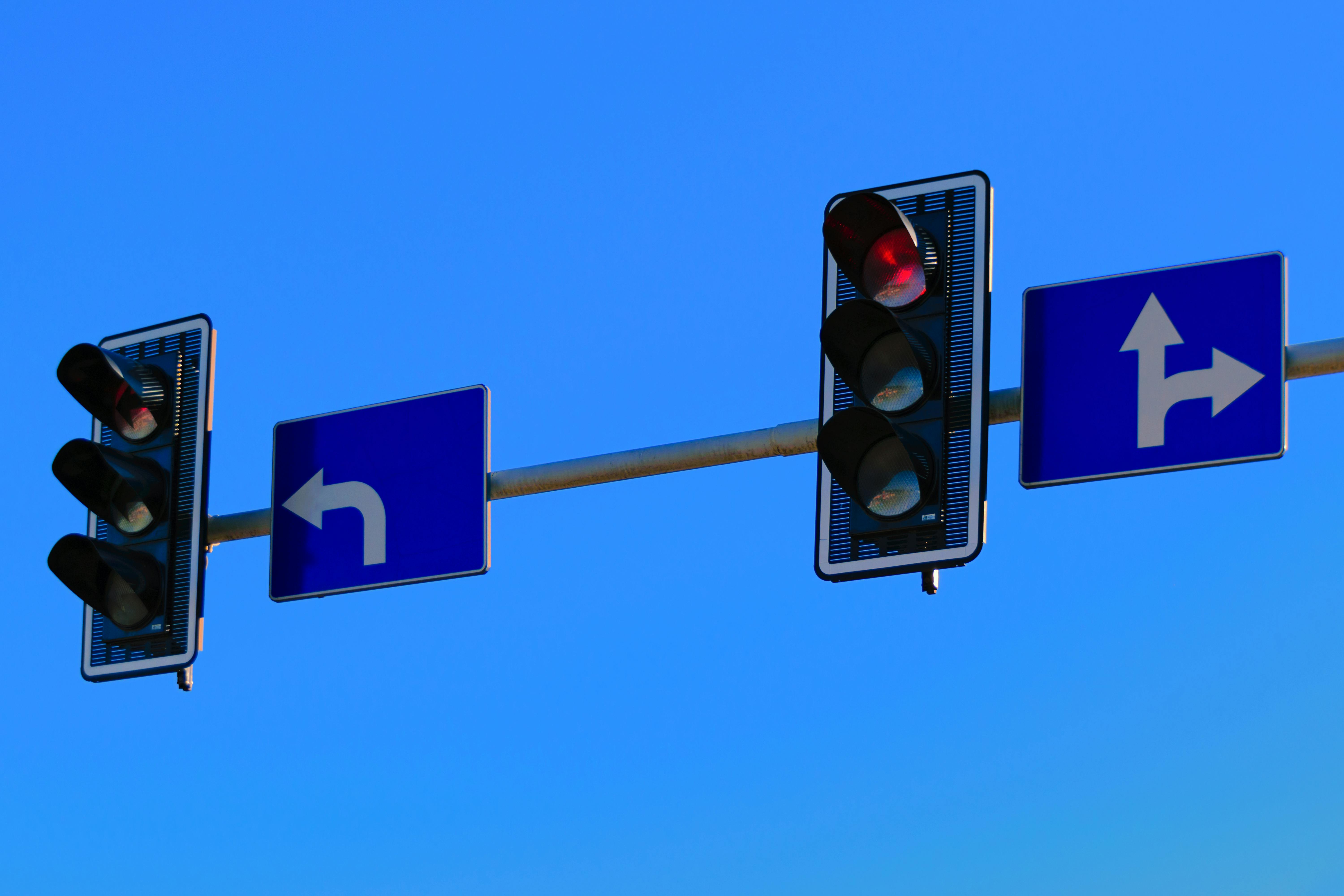 Traffic lights with road signs above an intersection. The intersection is not visible. The left road sign shows an arrow to indicate a left-turn lane. The right road sign shows to arrows to indicate a straight and right-turn lane.